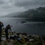 A dark, atmospheric and mysterious scene of Loch Ness in Scotland, shrouded in early morning mist. The vast, deep black waters of the lake stretch into the distance, framed by rugged Scottish Highlands and overcast grey skies. In the water, a subtle, ambiguous disturbance breaks the surface — a large dark shape or ripple suggesting something massive just beneath, neither clearly defined nor obviously explained. On the shoreline, a modern surveillance camera mounted on a pole and a small group of observers with binoculars and recording equipment hint at contemporary monitoring efforts. The overall mood is eerie, contemplative, and suspenseful. Cinematic editorial photography style, muted tones of deep green, slate grey, and black, with dramatic low diffused light filtering through clouds.
