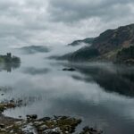 A misty, atmospheric scene at Loch Ness in the Scottish Highlands during early spring, captured in moody natural light. The dark, glassy surface of the vast lake reflects low-hanging grey clouds and rolling green hills. In the middle distance, a subtle dark elongated shape barely breaks the surface of the black water, ambiguous and mysterious — neither clearly an animal nor clearly an object. The shoreline is lined with ancient pine trees partially obscured by morning mist. The overall mood is eerie, contemplative, and cinematic, evoking both wonder and uncertainty. Photorealistic style with a cool, desaturated color palette dominated by deep greens, slate greys, and dark blues. Wide landscape composition, editorial photography aesthetic.