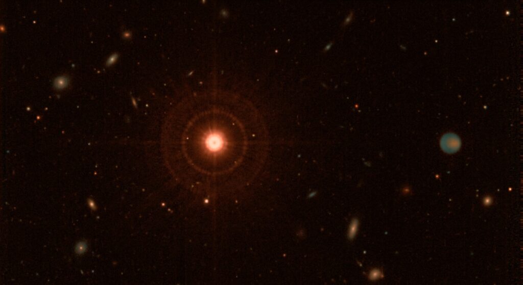 A dramatic deep-space scene showing a dim red dwarf star at center, glowing with a cool reddish-orange hue against the vast darkness of space, surrounded by a wide orbital region extending far into the outer reaches of the system. In the distant periphery, a massive Jupiter-like gas giant planet is visible, subtly illuminated by faint thermal infrared emission, its cold atmosphere swirling with muted bands of color at temperatures near -40 degrees Celsius. The scene is rendered in mid-infrared wavelengths, giving the image a warm thermal glow aesthetic with teal and amber tones representing heat signatures. A faint point spread function halo surrounds the central star, suggesting the challenge of direct imaging contrast. The composition emphasizes the vast empty space between the star and the distant giant planet, conveying the scale of tens of astronomical units. The background is filled with distant background stars and galaxies. The overall mood is scientific and awe-inspiring, rendered in a photorealistic space telescope imagery style reminiscent of James Webb Space Telescope observations.