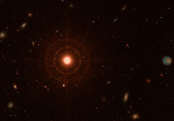 A dramatic deep-space scene showing a dim red dwarf star at center, glowing with a cool reddish-orange hue against the vast darkness of space, surrounded by a wide orbital region extending far into the outer reaches of the system. In the distant periphery, a massive Jupiter-like gas giant planet is visible, subtly illuminated by faint thermal infrared emission, its cold atmosphere swirling with muted bands of color at temperatures near -40 degrees Celsius. The scene is rendered in mid-infrared wavelengths, giving the image a warm thermal glow aesthetic with teal and amber tones representing heat signatures. A faint point spread function halo surrounds the central star, suggesting the challenge of direct imaging contrast. The composition emphasizes the vast empty space between the star and the distant giant planet, conveying the scale of tens of astronomical units. The background is filled with distant background stars and galaxies. The overall mood is scientific and awe-inspiring, rendered in a photorealistic space telescope imagery style reminiscent of James Webb Space Telescope observations.