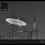 A high-contrast monochrome infrared surveillance still, styled as an authentic declassified military sensor capture: a smooth, elongated football-shaped object with three faint radial projections, glowing pale white-green against a grainy dark night sky, hovering above a distant horizon lined with the silhouettes of wind turbines. Subtle scan lines, sensor noise, and targeting reticle brackets overlay the frame, with a small telemetry readout box in one corner showing indistinct blurred numbers. The atmosphere is clinical and ambiguous rather than dramatic — no dramatic lighting, no lens flares, just the cold, flat realism of thermal camera footage. The composition should feel like a paused frame from an official military video, evoking mystery through technical starkness rather than spectacle, in a documentary photojournalistic style.