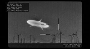 A high-contrast monochrome infrared surveillance still, styled as an authentic declassified military sensor capture: a smooth, elongated football-shaped object with three faint radial projections, glowing pale white-green against a grainy dark night sky, hovering above a distant horizon lined with the silhouettes of wind turbines. Subtle scan lines, sensor noise, and targeting reticle brackets overlay the frame, with a small telemetry readout box in one corner showing indistinct blurred numbers. The atmosphere is clinical and ambiguous rather than dramatic — no dramatic lighting, no lens flares, just the cold, flat realism of thermal camera footage. The composition should feel like a paused frame from an official military video, evoking mystery through technical starkness rather than spectacle, in a documentary photojournalistic style.