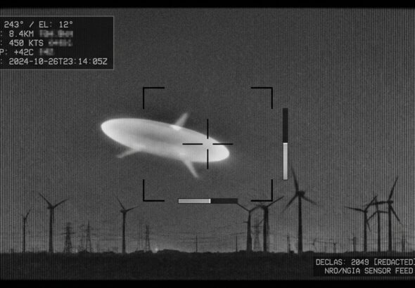 A high-contrast monochrome infrared surveillance still, styled as an authentic declassified military sensor capture: a smooth, elongated football-shaped object with three faint radial projections, glowing pale white-green against a grainy dark night sky, hovering above a distant horizon lined with the silhouettes of wind turbines. Subtle scan lines, sensor noise, and targeting reticle brackets overlay the frame, with a small telemetry readout box in one corner showing indistinct blurred numbers. The atmosphere is clinical and ambiguous rather than dramatic — no dramatic lighting, no lens flares, just the cold, flat realism of thermal camera footage. The composition should feel like a paused frame from an official military video, evoking mystery through technical starkness rather than spectacle, in a documentary photojournalistic style.